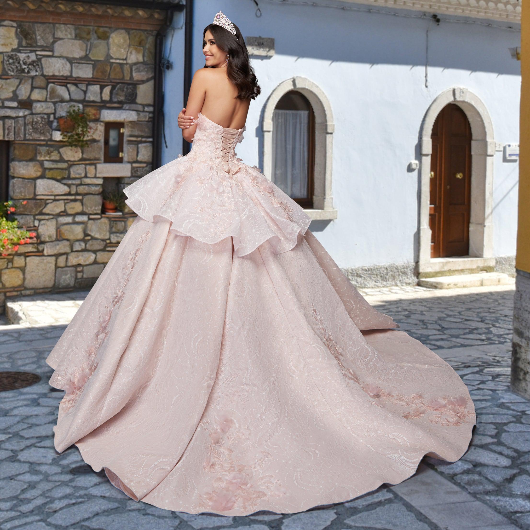 A woman in the Quinceañera Sample Dress QF115—a strapless, pale pink ball gown with a tiara—stands on a cobblestone street before charming stone and stucco buildings.